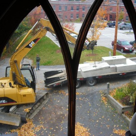 Man in street with arms folded watching excavator lift concrete blocks from trailer.